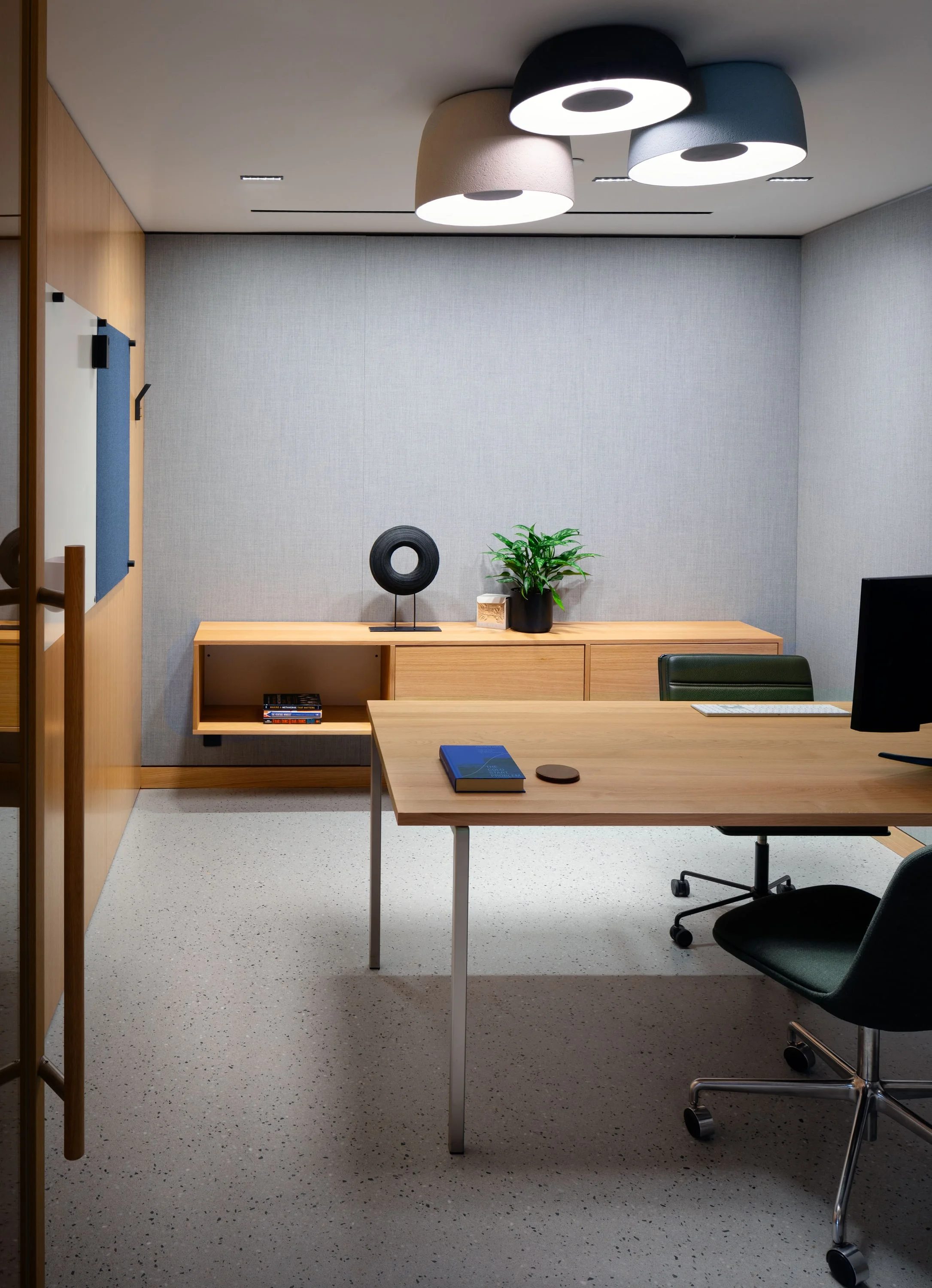 Büro mit Holzschreibtisch, Wandregal aus Eiche, schwarzen Bürostühlen und runden Deckenleuchten.
