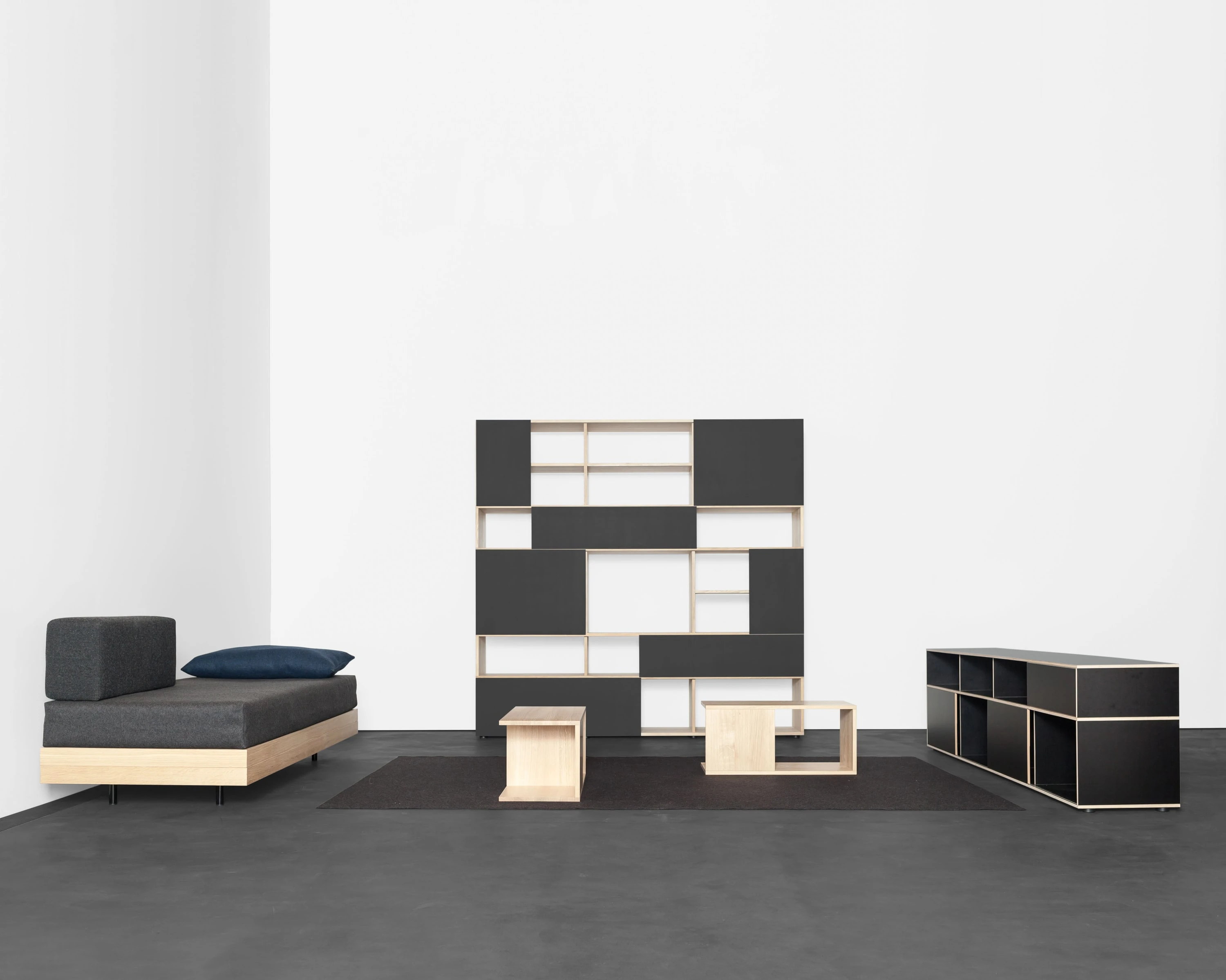 Raum mit Tagesbett und dunklen Kissen, schwarz-weissem Regal, kleinen Holzkuben auf dunklem Teppich und schwarzem Sideboard.