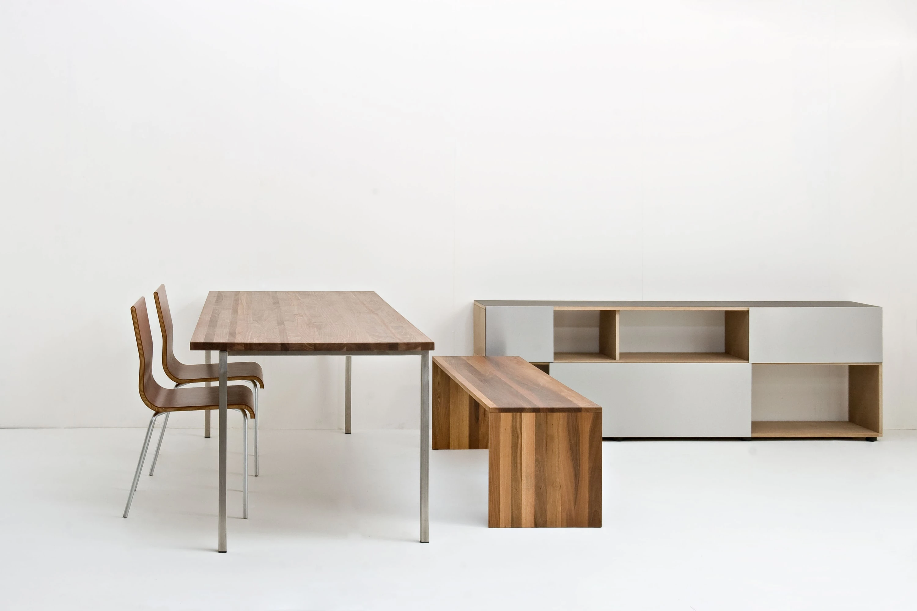 Holztisch auf Stahlbeinen mit zwei Holzstühlen und einer Bank aus dunklem Holz, dahinter ein Sideboard mit hellgrauen Fronten.