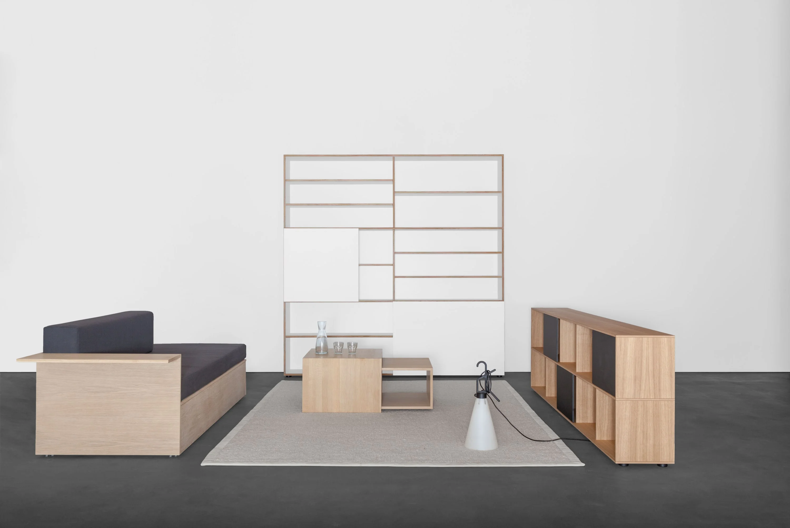 Couch aus Eiche mit dunkler Auflage, hohes weisses Regal, kleine Holzkuben auf hellem Teppich und ein Sideboard mit schwarzen Fronten.