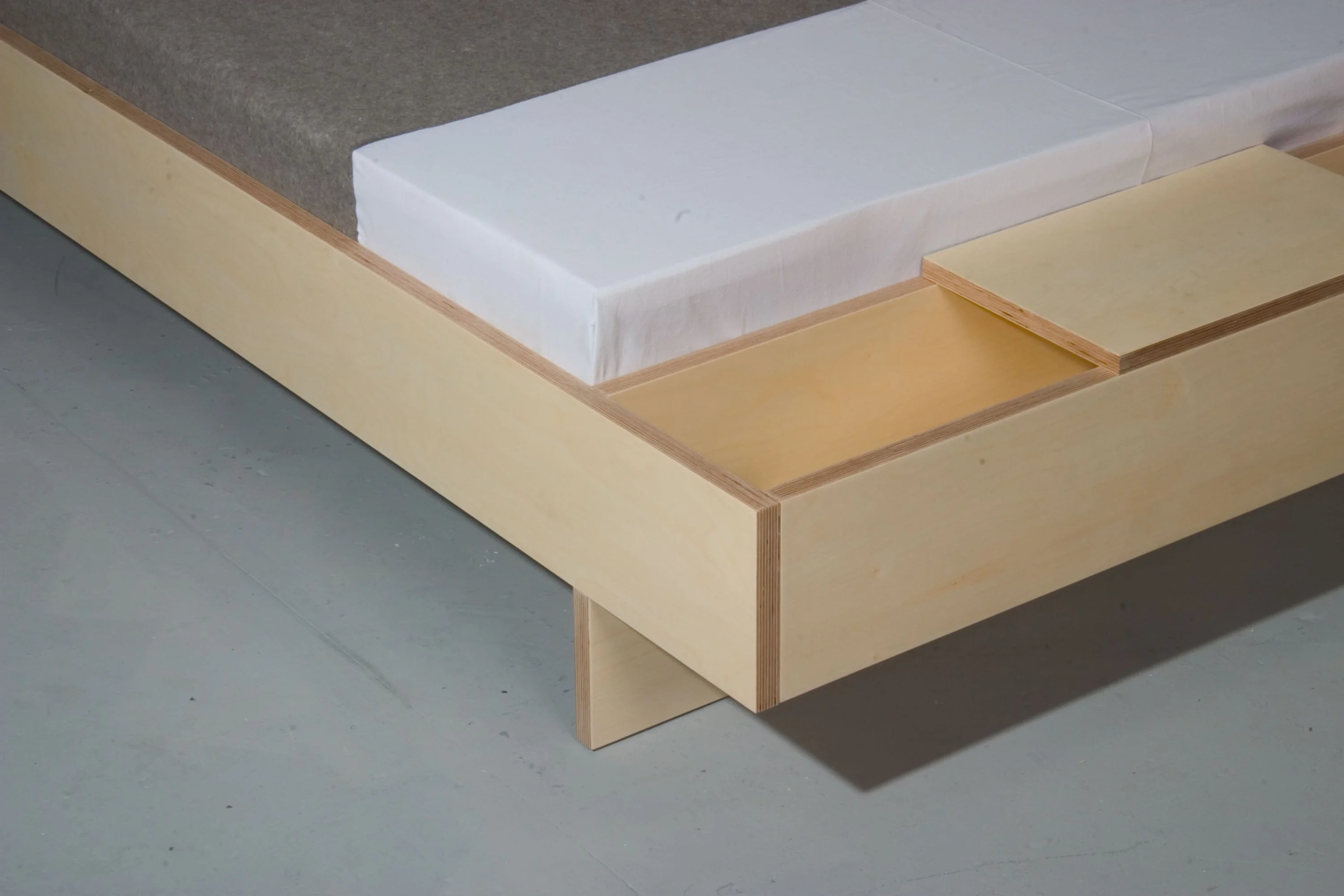 Nahaufnahme einer Bettecke aus hellem Holz mit geöffnetem Staufach neben der Matratze.