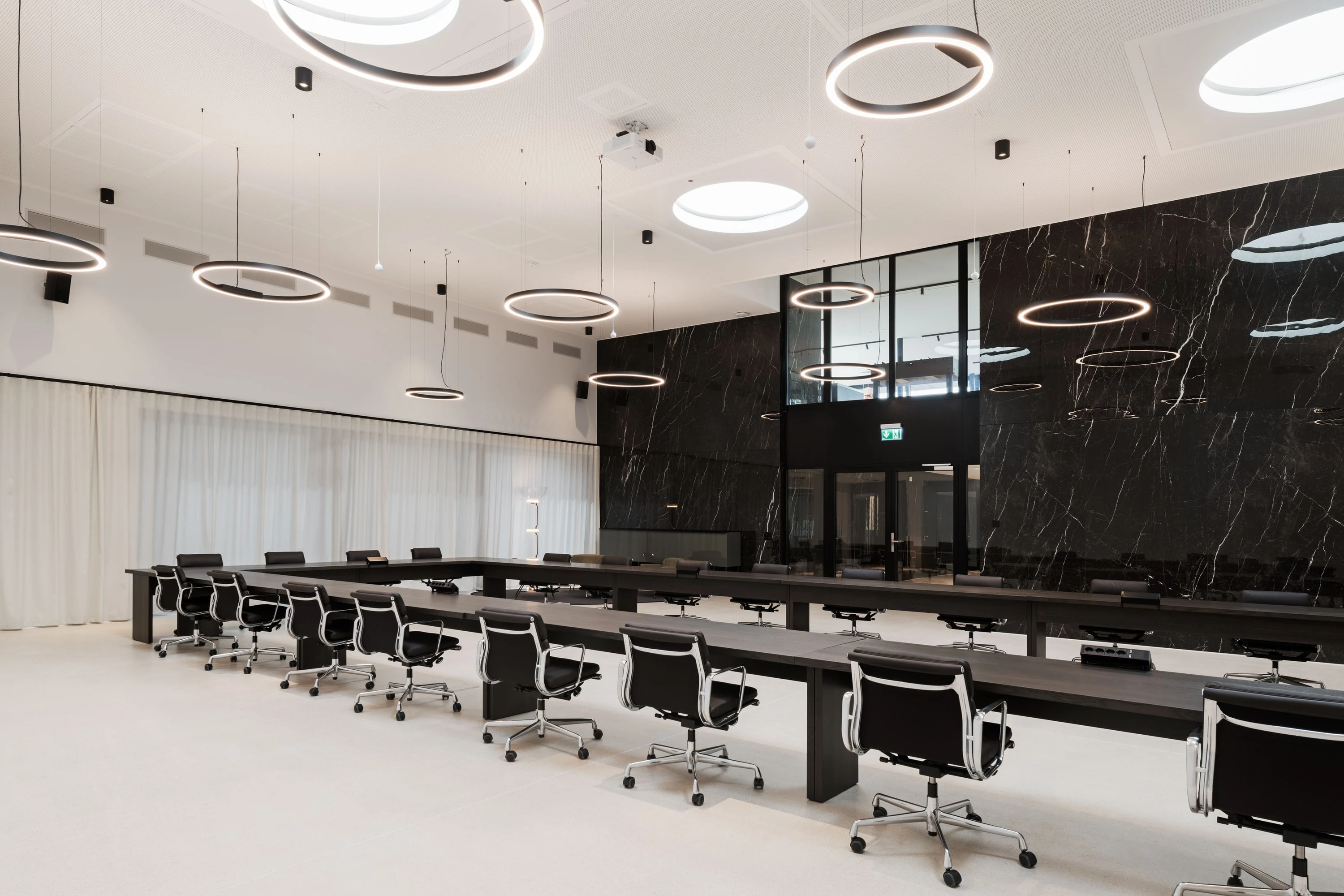 Grosser Konferenzsaal mit langen schwarzen Tischreihen, Bürostühlen, Ringleuchten und dunkler Marmorwand.