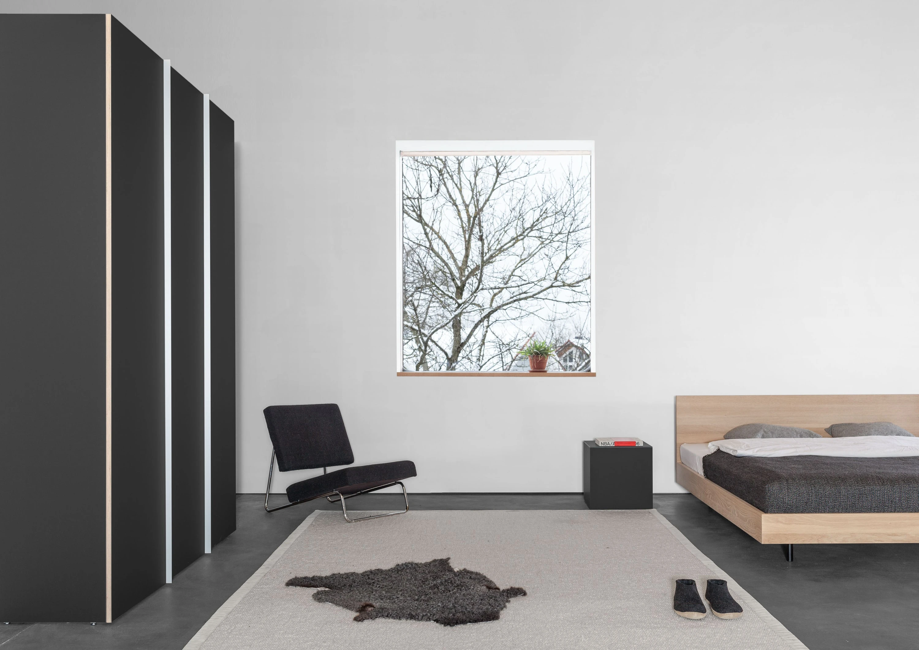 Schlafzimmer mit hohem schwarzem Schrank, Fenster, schwarzem Liegesessel, Hocker und Bett aus Eiche mit hohem Kopfteil.