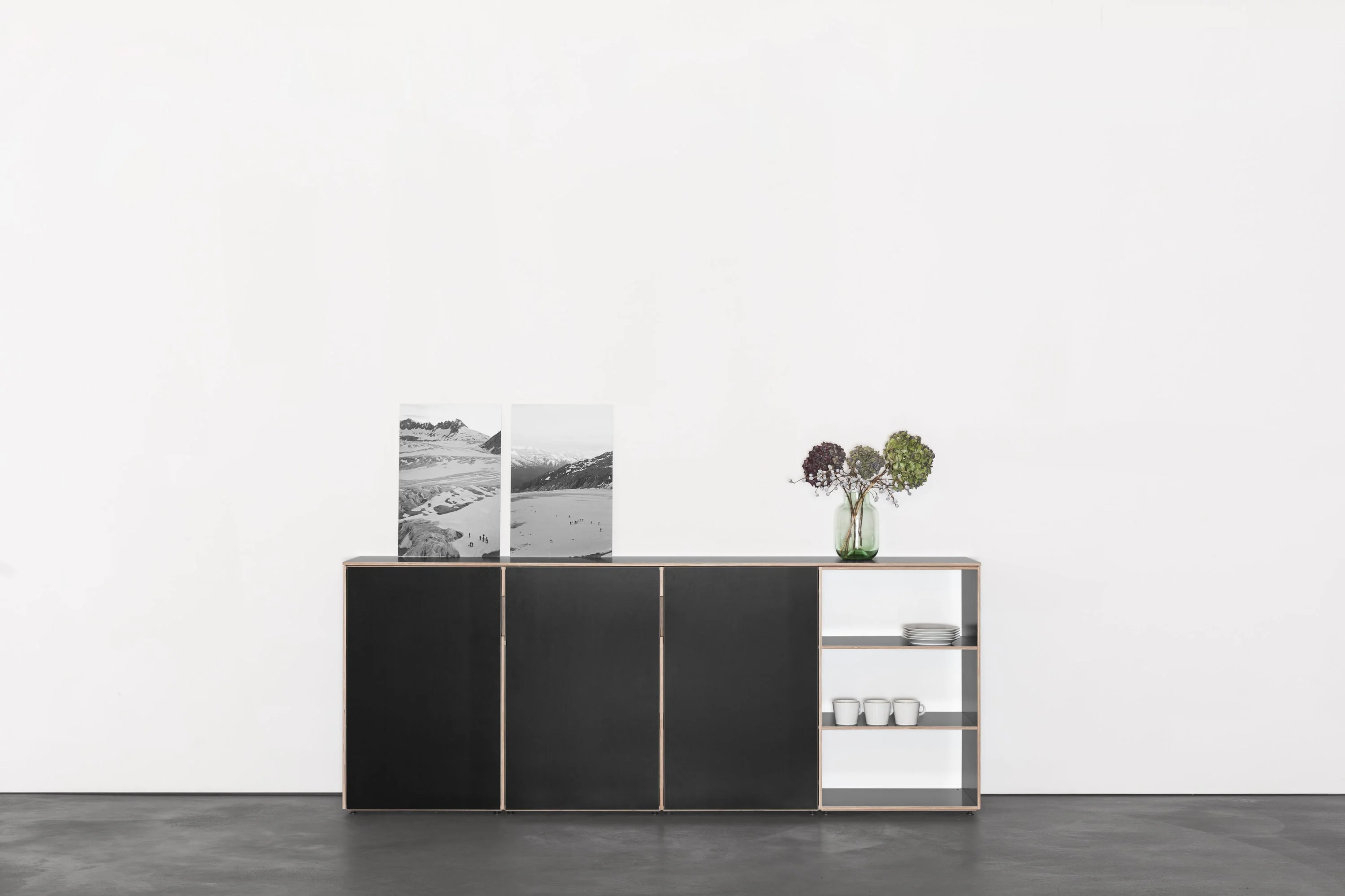 Schwarzes Highboard mit offenem Fach, darauf zwei Fotografien und Blumen.